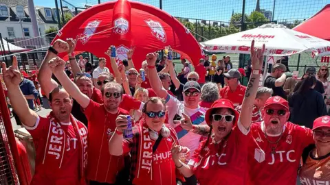 Jersey Bulls Supporters Group Jersey Bulls fans smile and raise their hands at the camera at the fan zone