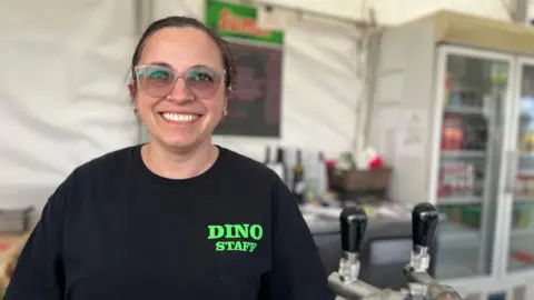 Anita smiles at the camera at a food stall as she is next to some beer taps. She has a black t-shirt on which says Dino Staff and her hair is tied into a ponytail. She has clear frame glasses on as well.