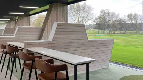 A golfing green is outside with a table and chars inside a driving range , with a large barrier between ranges under cover.