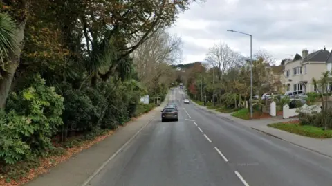 A picture of the street view in Falmouth. There is a large road with a few cars on it and houses to the right of the image. To the left, there is trees.