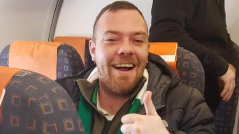 Kevin Davidson, smiling at camera on a chair on a plane with his thumbs up 