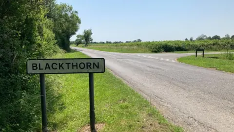 A rural road with a Blackthorn sign to the left. There are no cars.