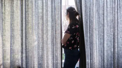 A woman looking out a window in between partially drawn curtains with her arms folded. 