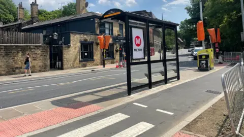 Sofía Luis-Hobbs/BBC A bus stop is pictured on an island in front of a cycle track. There is a pub on the other side of the road. There is greenery around on either side of the road.