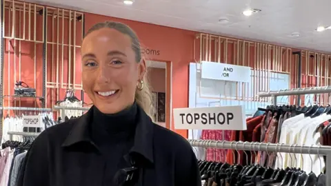 Alicia Owen/BBC A young woman with her blonde hair tied back in a ponytail. She is wearing a black rollneck jumper with a black jacket over the top. Clothing rails are behind her.
