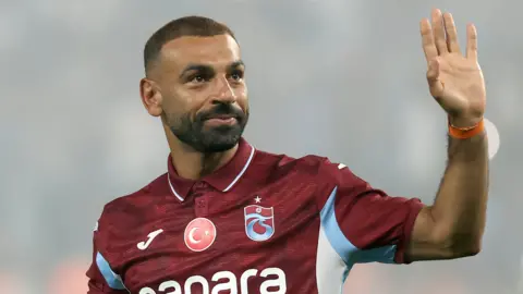 Mohamed Salah, visible from chest up wearing a claret Trabzonspor polo shirt with white and sky blue trim and a circular Turkish flag in the upper centre of the chest, raises his left hand to wave while showing a closed-mouth smile on his face