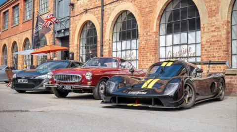 Lewis Warren Three cars parked outside a brick industrial-style building with large arched windows. In the foreground is a low black race-style car with yellow stripes; behind it are a red classic coupe and a dark modern sports car. A Union Jack flag and outdoor seating with umbrellas are visible.