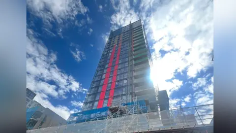 Craig Robinson The outside view of the large tower block, with the sun shining, and scaffolding visible all over 