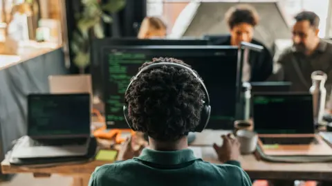 A person, pictured from behind, sitting at a desk. There is a computer and two laptops in front of them. They have short brown hair and are wearing a dark green shirt and black headphones. 