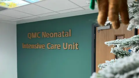 The entrance to the new unit with the words QMC Neonatal Intensive Care Unit embossed on a green wall. Part of a Christmas tree and a brown door is pictured to the right of the picture