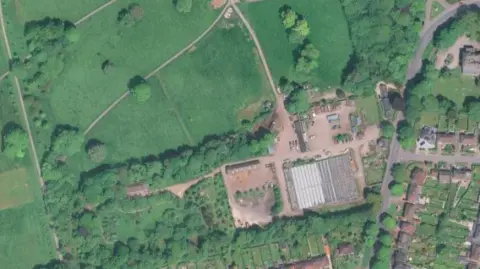 Google Satellite image showing large swathes of grassland with some woodland areas. A non-grassed area in the centre shows cars parked and what looks like a large greenhouse.