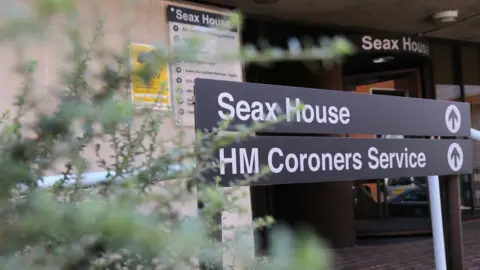 A sign at the entrance to Seax House, which is home to the coroner service. The sign is black and has the words "Seax House" and "HM Coroners Service" in white font. There is a bush to the left of the sign, with a revolving door entrance behind it.