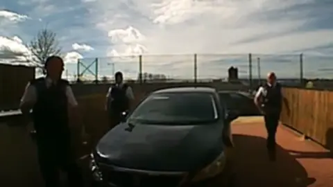 Jacqueline Cowdall A still image from a doorbell camera showing three male police officers in black uniforms walking on a driveway, where a black car is parked.