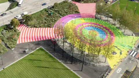 Andy Stagg A picture of colourful patterns drawn on the pavement and the square outside Milton Keynes station. There are yellow and pink circles on the square. The walkways leading to it are painted in red-and-white stripes.