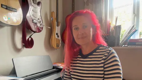 BBC Sharon Stanton is looking into the camera while sat at a piano, with some guitars on the wall behind her. She has long red hair and is wearing a blue and white top.