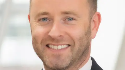 Handout A man with a short stubble and wearing a black suit and white shirt smiling into the camera.