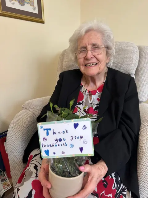 Care UK A lady with short white hair and glasses, sat on a cream coloured chair, smiles as she holds a pot of flowers with a note saying 'Thank you from Penn Fields'.