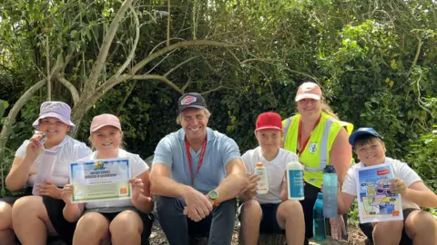 Charlotte, 10, Hattie, 9, Tony Tostevin (MUG), Ollie, 10, Juliet Bell (Vale Primary School Sun Safety Ambassador teacher) and Lewis, 9, sat in a shaded woodland area at the school. They are all smiling, wearing sun hats and holding an array of items like suncream, the sun-safe accreditation certificate and waterbotles. 