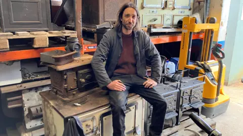 Olly Osborne in work clothes, sitting in his workshop on an old metal cooking range. There are a variety of other historic ovens to his side and behind him. He has long hair and a short beard. 