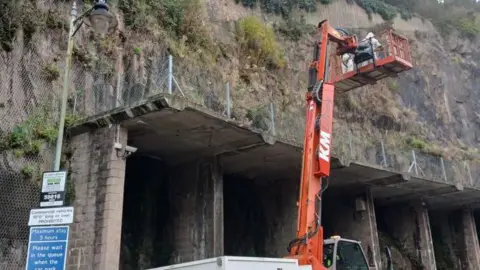 Jersey Asian Hornet Group A cliff face is seen with a crane in front of it, on which two people stand in white protective suits in preparation for removing an Asian hornet nest, while in the foreground a lamp post and signage can be seen. 