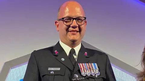bald man wearing glasses and dark grey uniform with medals is smiling