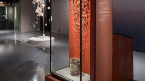 Items from an exhibition at Jersey Museum about the story of the island. The item at the front of the image is an old pot encased in a glass box with an orange display behind it. Pieces of pottery are on the wall of the display.