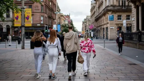 Four young people are walking away from the camera down the centre of the street. They are dressed casually, wearing hoodies, jogging bottoms and trainers. One person is carrying a brown bag, and another has a small backpack. They are on a pedestrian-friendly street lined with historic buildings featuring ornate architecture