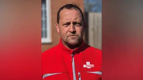 Stephen Harvey A postman wearing a Royal Mail top. He is looking at the camera. He has short black hair and has stubble on his face. There is a red-brick building and a wooden fence behind him.
