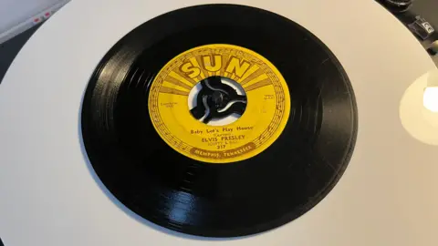 John Devine/BBC An original Sun Records 45 rpm vinyl record on a turntable. The label on the disc is yellow background with light brown writing it is a song called "Baby Let's Play House" from 1955 and was recorded by Elvis Presley at the Sun studio in Memphis.