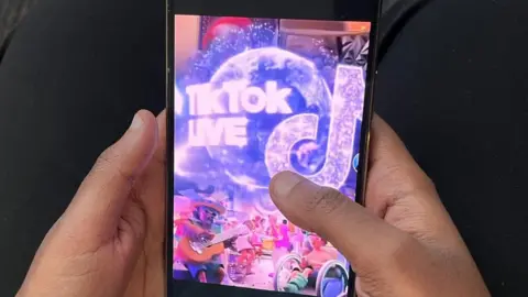 BBC Muestra la mano de alguien sosteniendo un teléfono móvil con TikTok Live en la pantalla.