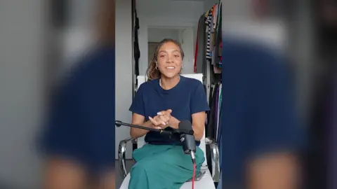 Miquita has tied-back hair and is wearing a blue t-shirt and green trousers sat on a white seat