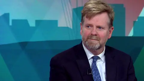 Head and shoulder shot of Nick Oliver short light brown hair and a trimmed beard, streaked with grey. He is wearing a dark jacked, white shirt and a blue tie and seated in front of a background featuring different shades of blue.