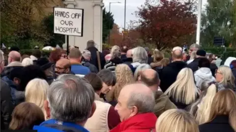 Steve Beresford Many people, men and women, gathered outside, surounded by trees. One person holding a placard that reads "hands off our hospital". 