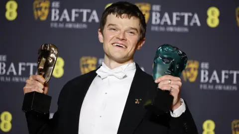 EPA/Shutterstock Robert Aramayo, a young man with short dark hair, smiling broadly on the red carpet at the Baftas. He is wearing a tuxedo with a white bowtie and holding up two face-shaped Bafta awards, one gold and one green. 