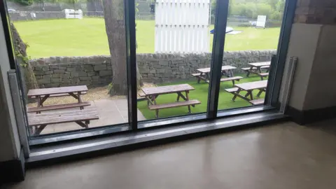A window with white brackets round the edge. Through the window several picnic benches can be seen outside.