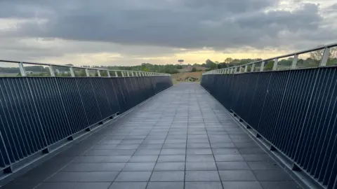 A view from the apex of a footbridge, looking towards countryside in the distance. There are silver-coloured railings on either side and the path is pathed in square tiles. 