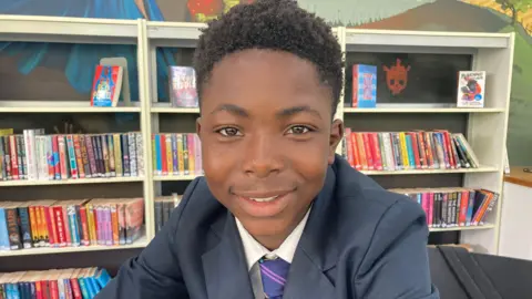 BBC/Carla Fowler Star seated indoors in what appears to be a school library or study area. He is positioned centrally in the foreground and is leaning slightly forward toward the camera, with one arm resting across the front of the body. He is wearing a dark blazer over a white shirt and a striped tie with diagonal bands in multiple colors, suggesting a school uniform.