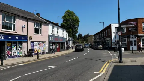 The centre of Abbots Langley, showing a road junction with shops to the left and right. A tree is visible in the background. There are double yellow lines on the side of the road to the right.