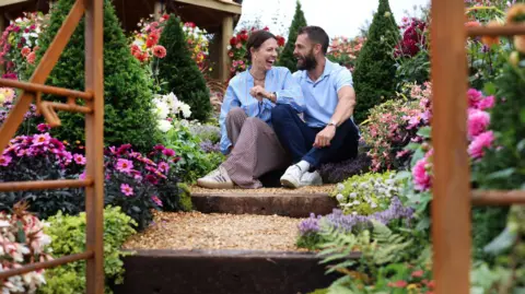 Gareth Jones Photography A couple pictured sitting on a step in a garden, laughing, surrounded by flowers.