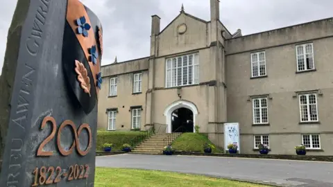 The historic stone Lampeter campus of the University of Wales Trinity St David building, with stone steps leading up to a large arched entrance. There are multiple mullioned windows. A slate sign in front of the building has copper coloured lettering which says '200' and '1822-2022'.