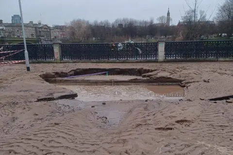 Scottish Water Damage caused by a burst 24-inch (60cm) main water pipe on Florence Street, Glasgow. A road has a wide flooded hole and the surrounding carriageway and pavements are covered in rippled mud. Red and white cordon tape is tied to a lamp post. A fence along the river bank is in the background, with a view of Glasgow Green in the distance.