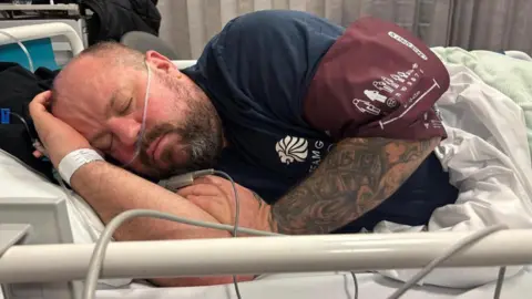 Russ Lee Russ Lee lying in hospital bed asleep wearing a navy t shirt. He is a wearing a hospital wrist hand on his left wrist and hs a tube 