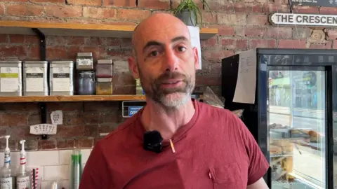 BBC/NATHAN TURVEY Tim Linley, who has a grey and brown beard and is wearing a read t-shirt, standing in his coffee shop. There are coffee tins on a shelf behind him and a fridge.