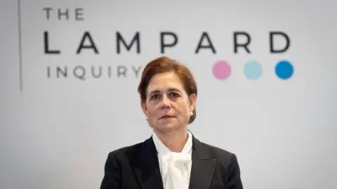 PA Media Baroness Lampard looks at the camera. She is in a black jacket and white blouse with a bow at the front. She has short brown hair in a bun and pearl earrings, she is standing in front of a Lampard Inquiry banner