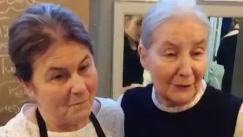 Two women stood side by side wearing aprons or a tabard speak to a reporter in a busy cafe