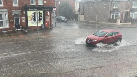Heavy rain hits the road with a red car driving through it. In the background is a Premier corner shop with black signage. 