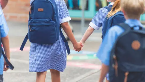 Getty Images Pictured from behind, two young girls in blue check-pattered dresses walk towards a school, hand-in-hand. A boy with a backpack is also pictured, in a short-sleeved blue shirt. 