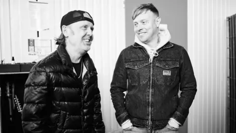 Ed Mason A black and white photo of Sam Carter and Metallica drummer Lars Ulrich. Sam's standing with his hands in his pockets, looking fondly at Lars. Lars is perched on some sort of box or equipment, and smiling. He's wearing a black puffer jacket and backwards baseball cap. They appear to be in a backstage area and the mood is relaxed.