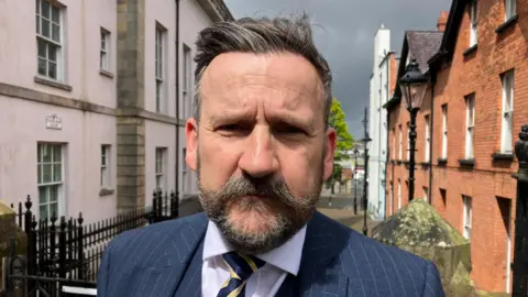 Derwin Harvey has dark hair flecked with grey and a dark beard and moustache. He is wearing a blue pinstripe suit with a white shirt and a navy and gold striped tie. He is standing in a street.