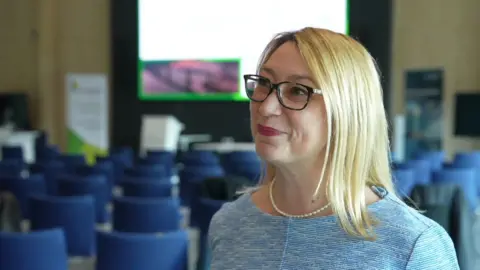 Jess Asato standing at the back of a conference room with rows of blue chairs, which are currently empty. There is a large white screen at the front of the room behind Jess. She has black glasses, pink lipstick and blonde hair.